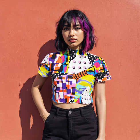 A colorful 90s-style mesh crop top featuring a maximalist abstract pattern. This see-through shirt has a high mock neck, short sleeves, and a vibrant, retro design with geometric shapes, color blocking, and polka dots in bold shades of pink, yellow, blue, and orange. The Y2K-inspired top is a statement piece of festival clothing, perfect for a rave outfit or alternative, dopamine-dressing-inspired streetwear looks.